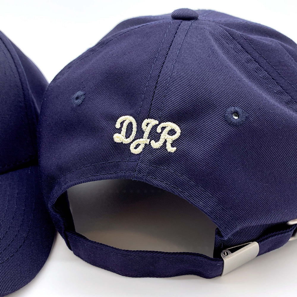 image of the back of a navy cap with initials embroidered in a script font using cream thread.